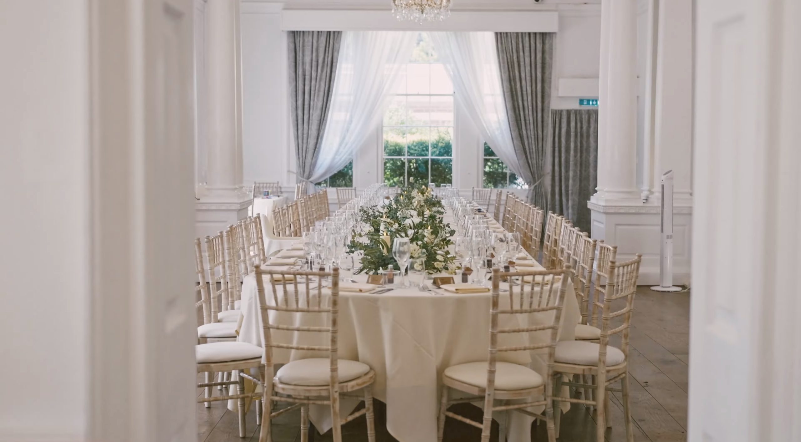 Elegant banquet table set with white cloth, greenery centerpiece, and gold chairs in a bright hall with tall windows and curtains at Bawtry Hall