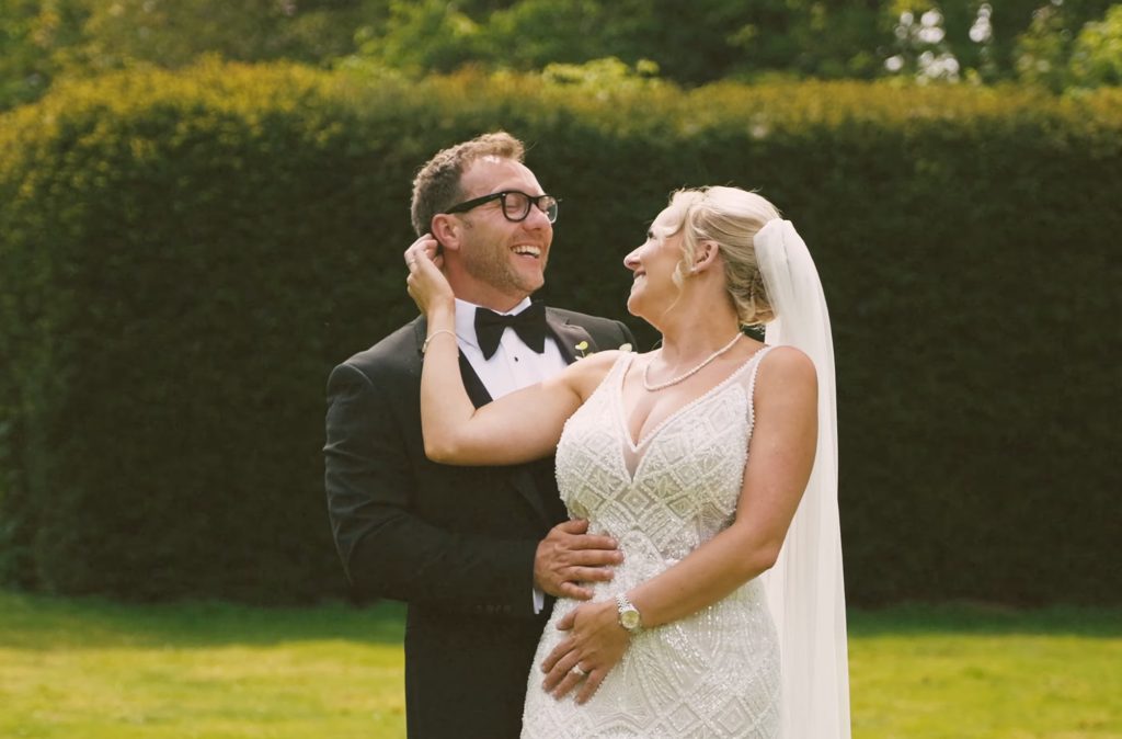Smiling bride and groom in wedding attire embrace on a sunny lawn with a dark green hedge background, at Bawtry Hall