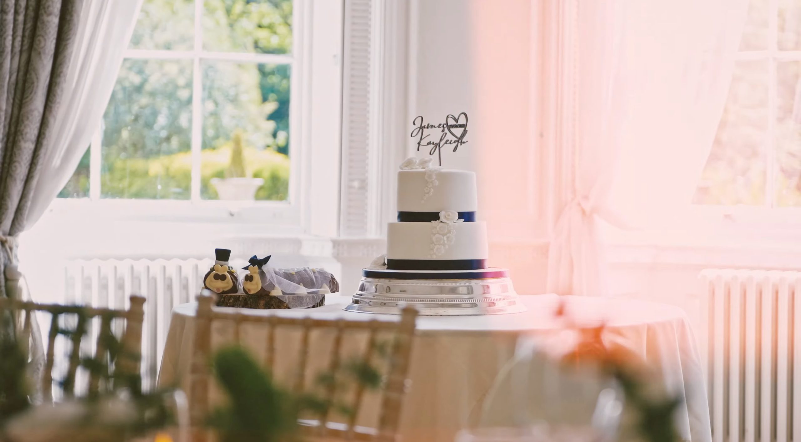 White wedding cake on a table by a bright window, with James and Kayleigh topper and small figurines nearby,  at Bawtry Hall